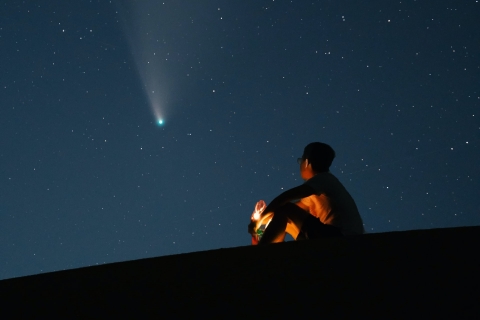 Man sitting on hill gazing up at green-headed comet flying in night sky