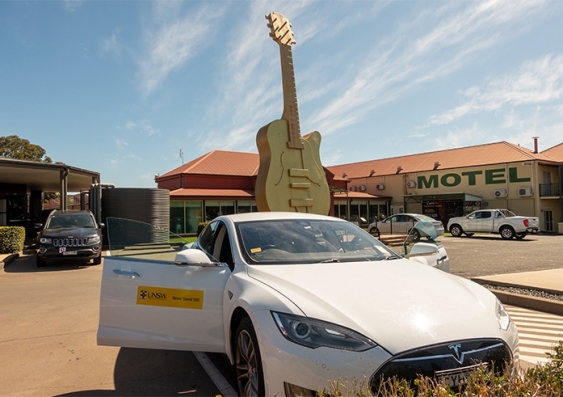 A Tesla car with UNSW emblem in front of The Big Golden Guitar, Tamworth