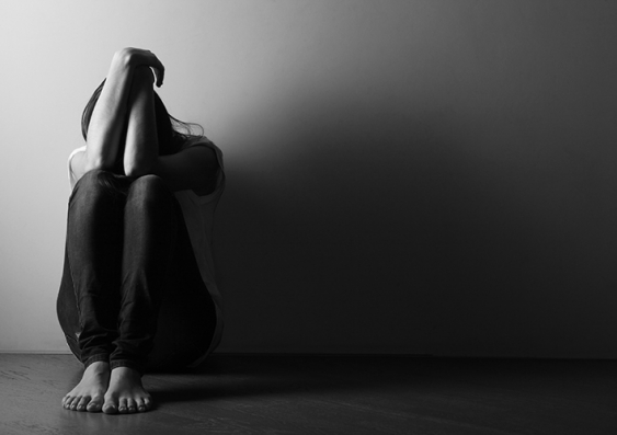 A person sitting down against the wall in a darkened room hiding their head in their hands