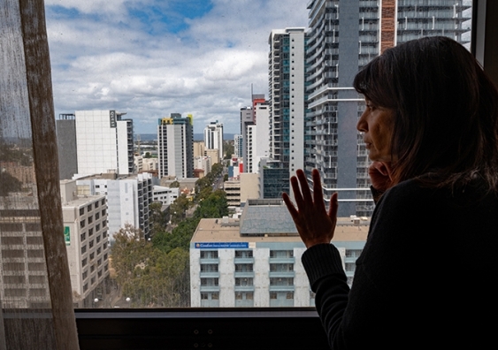 A woman peers out at the city from a hotel room some floors up