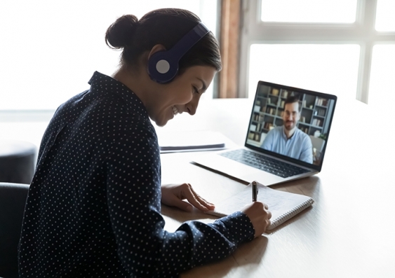 A smiling woman wearing headphones takes notes from an online lecture viewed on her laptop