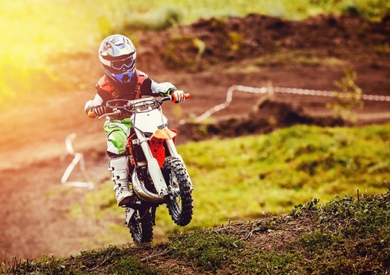 A child wearing helmet and motorcycling gear does a wheelie on a dirt track