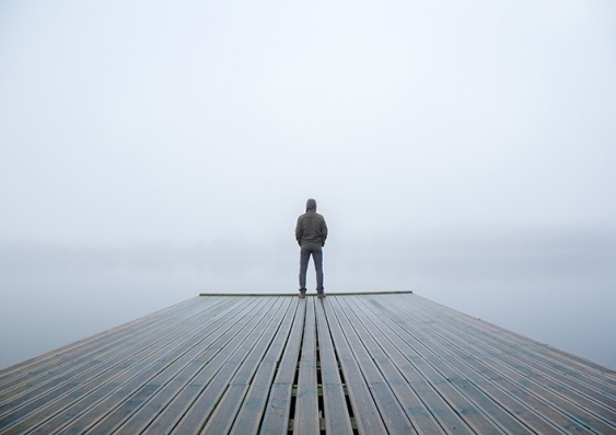 A lone figure in a hoodie stands at the end of a wharf peering into the mist