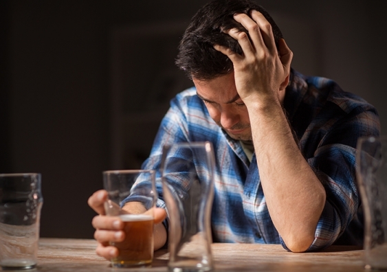 A man wearing a flannel shirt holds his head in one hand, the other hand holding a beer