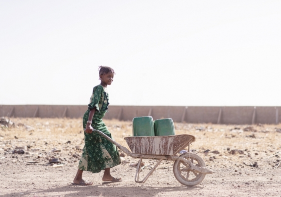 A young African girl carries buckets of water in a wheelbarrow across drought-stricken land.