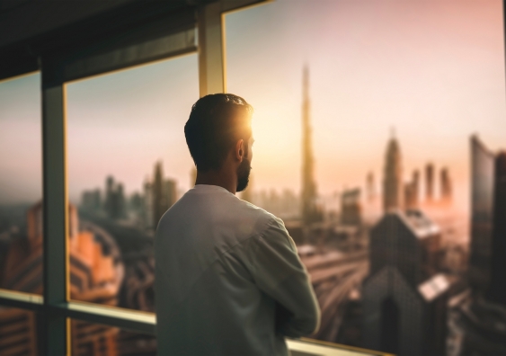 behind view of a man looking out a window towards a city at sunset