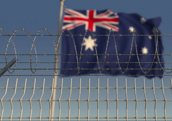 A blurred Australian flag waving behind a barbed wire fence. Image: Shutterstock