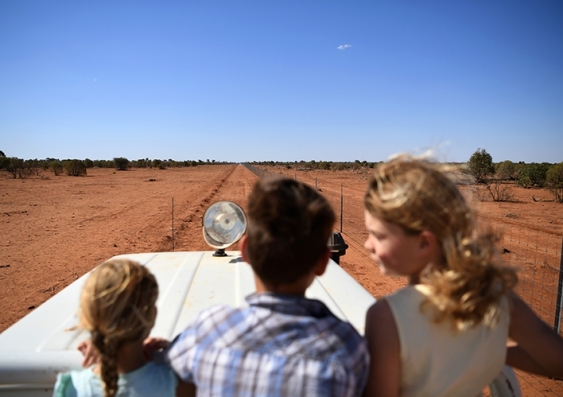 Three kids on the back of a ute travelling down a long stretch of dirt road in central Australia