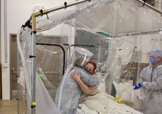 A patient in bed receiving a hug from a clinician in the Care Cube, an airborne infection isolation tent