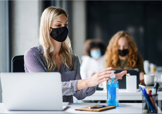 Woman sitting at desk in workplace with mask on using hand sanitizer. 