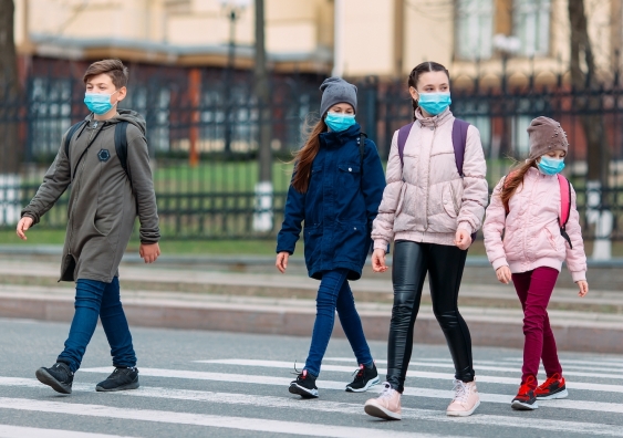primary school-aged children walk across a zebra crossing wearing face masks