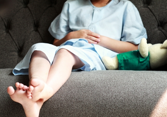 legs of patient kid in blue patient suit sitting on sofa and touch or press on her abdomen with her doll. 