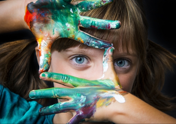 a young woman stares at camera through her fingers which are covered in colourful paint
