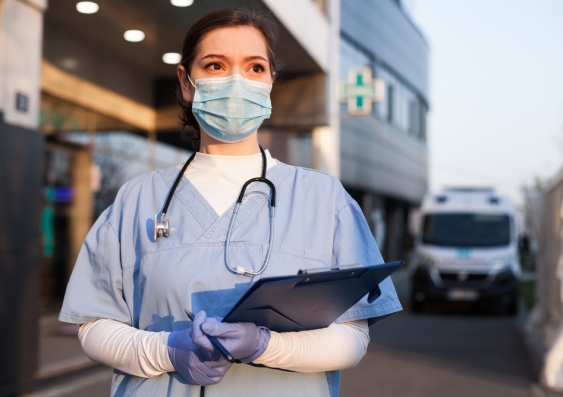 Young female EMS key worker doctor in front of healthcare ICU facility, wearing protective PPE face mask equipment, holding medical lab patient health check form