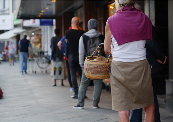 People standing a metre from each other outside of a shop with their backs turned from the photograph. A woman holding her wicker basket is the focus.
