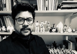 Dr Eduardo Benitez Sandoval sitting in front of shelves of robots, figurines and books