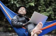 A contented man wearing glasses and a cap lies in a hammock with a laptop