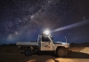 A white ute with 'Wild Deserts' logo under a starry night sky. Reece Pedler shines a spotlight from the ute into the distance.