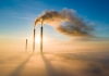 Waste gas is shown escaping two industrial chimneys against a blue sky background and surrounded by fog