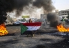 A man holding a Sudanese flag kneels and looks skyward among burning tyres 