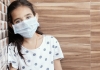A young girl wearing surgical mask leans against a wall