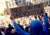 A hooded figure holds up a placard with "nice day for a revolution" during a protest