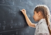 A young girl writes on a chalkboard with maths equations on it