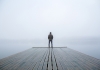A lone figure in a hoodie stands at the end of a wharf peering into the mist
