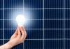 a hand holds a lit lightbulb in front of a solar panel