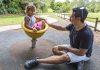 an indigenous child sits on play equipment smiling accompanied by an adult