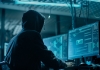A man wearing a hood looking at computer screens. 