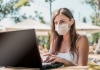 Woman wears face mask while sitting at a table by the beach with her laptop