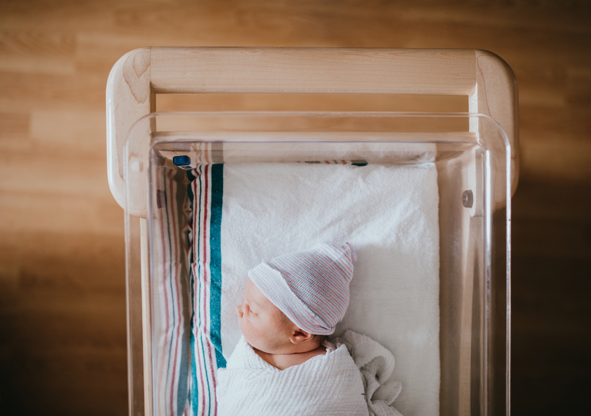 A baby just born at the hospital rests in a hospital bassinet crib, wrapped in a swaddle and wearing a beanie hat.