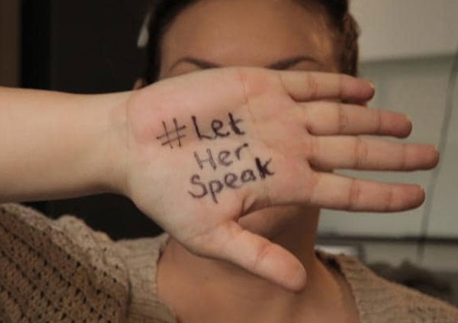 Woman holding her hand in front of her face with the words #Let her speak written on her palm